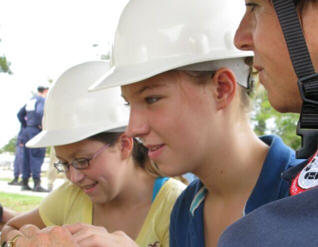 Woman showing young women how to do a technical task wearing hard hats.